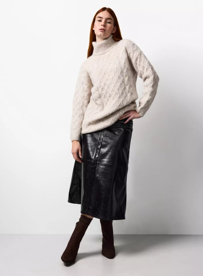 A woman models a beige turtleneck sweater with a long black leather skirt and brown boots.