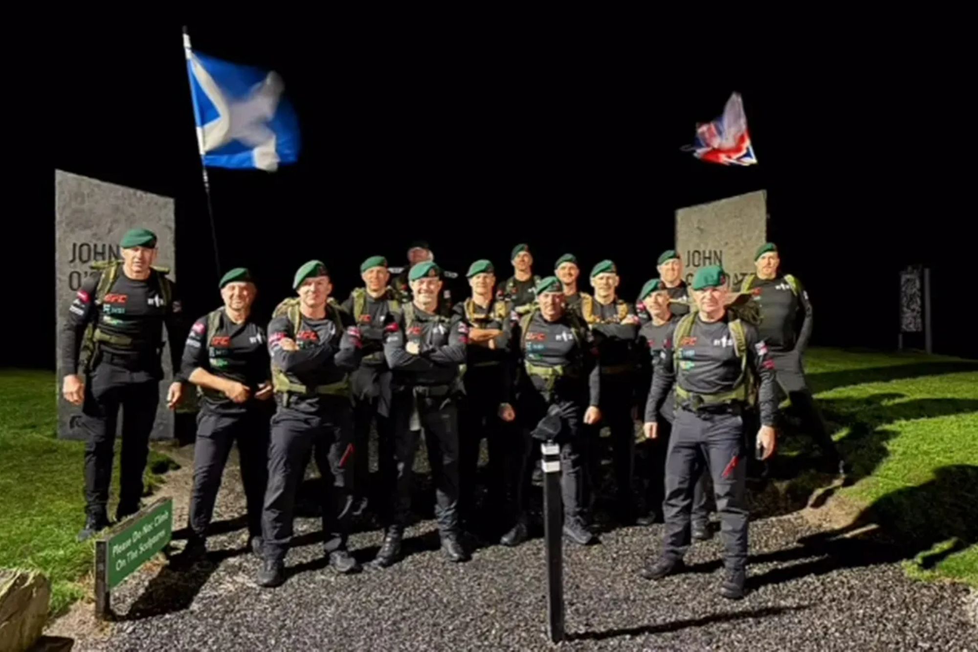 An image collage containing 1 images, Image 1 shows A group of 15 men, some with backpacks, stand together at night in front of two large stone markers and a Scottish flag