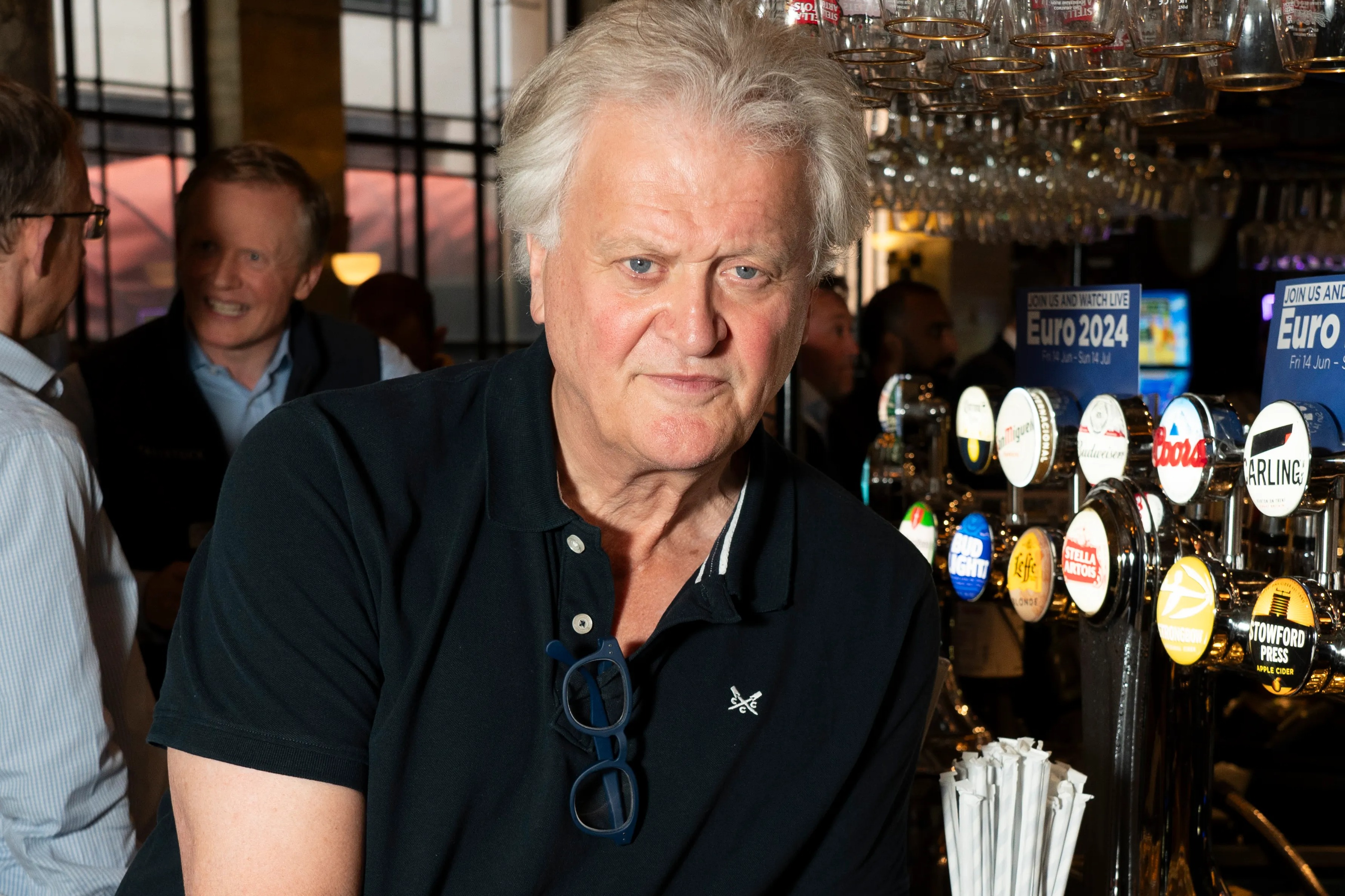 Tim Martin leaning on a bar with a pint of beer in hand, surrounded by beer taps and a "Euro 2024" sign.