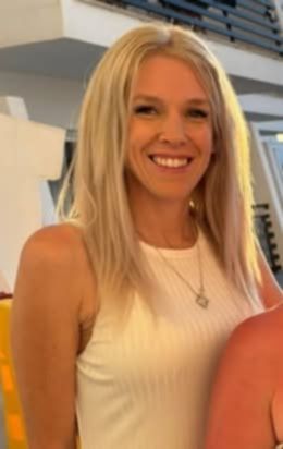A young woman with long blonde hair smiles while wearing a white ribbed tank top and a silver necklace.