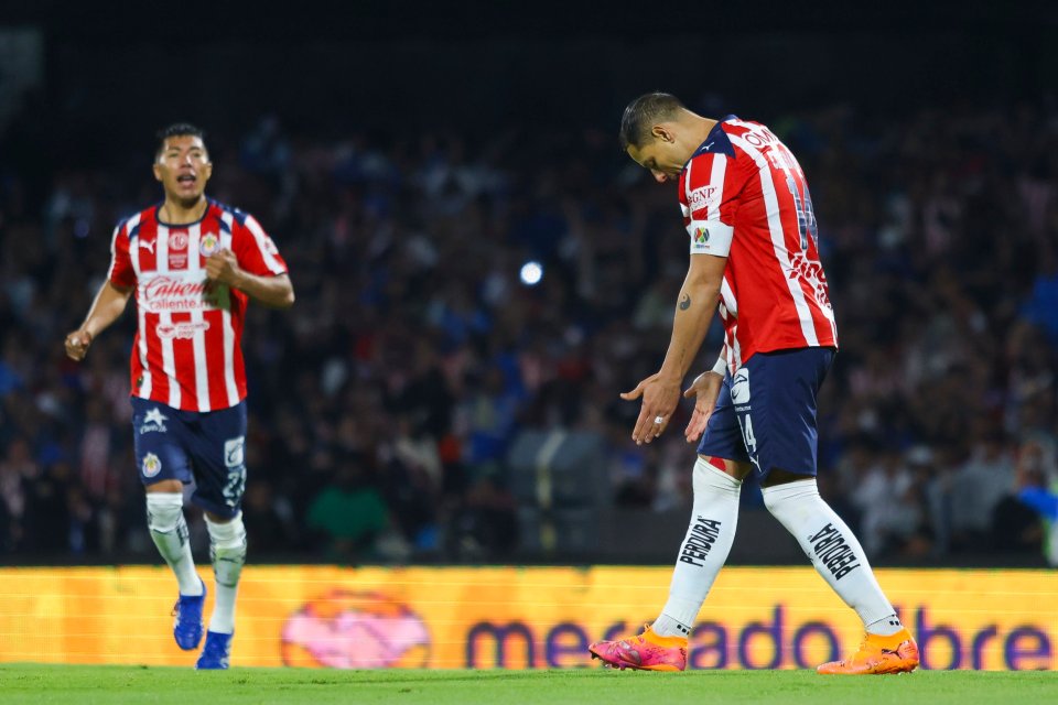 A Chivas player in a red and white striped jersey and navy shorts looks dejected, hands out, while another Chivas player in the background cheers.