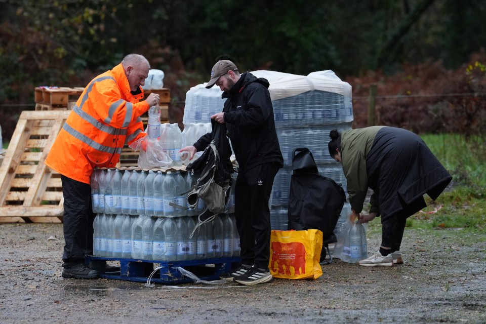 A worker in an orange high-visibility jacket hands out bottled water to people from a stack of palettes.