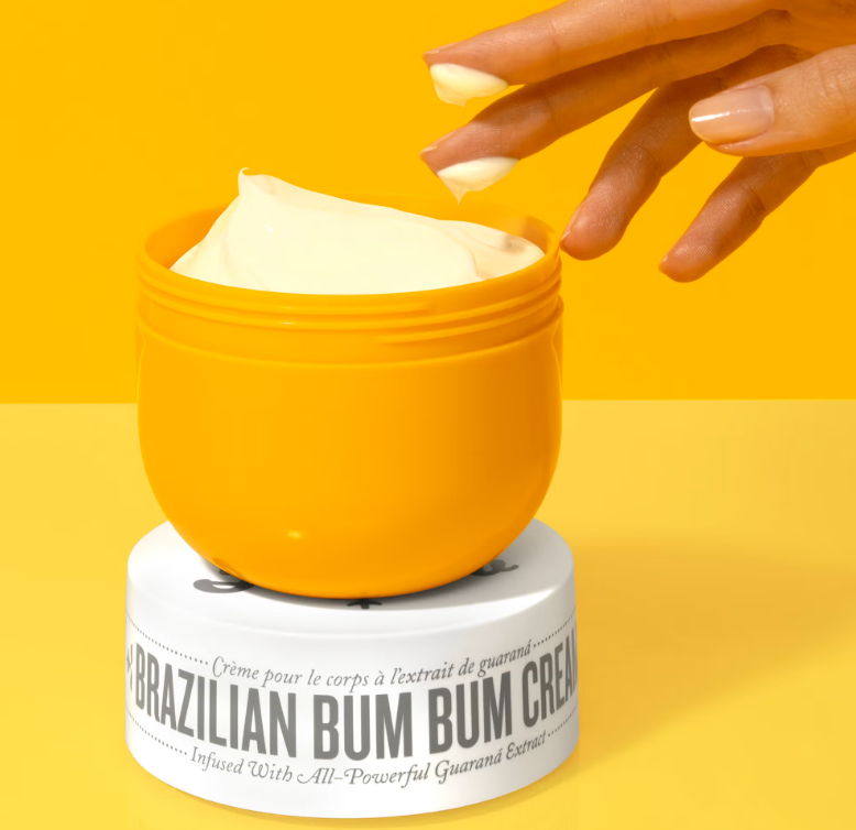 A hand with cream on two fingers above an open jar of Sol de Janeiro Brazilian Bum Bum Cream.