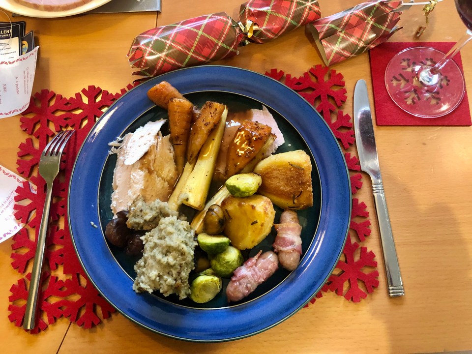 A plate of Christmas dinner with turkey, carrots, parsnips, roast potatoes, sprouts, pigs in blankets, and stuffing, surrounded by Christmas crackers and red snowflake placemats.