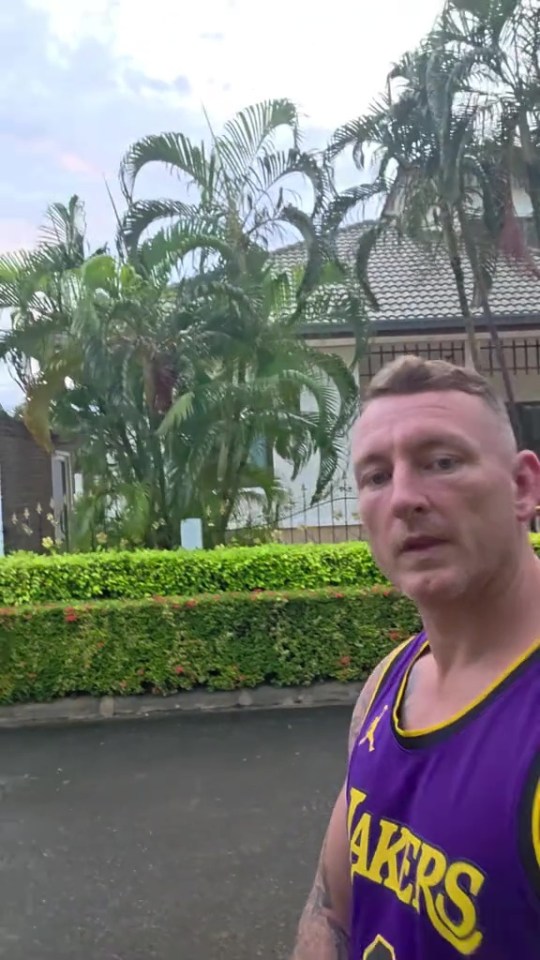 A man in a purple Lakers jersey standing on a street in front of houses and palm trees.