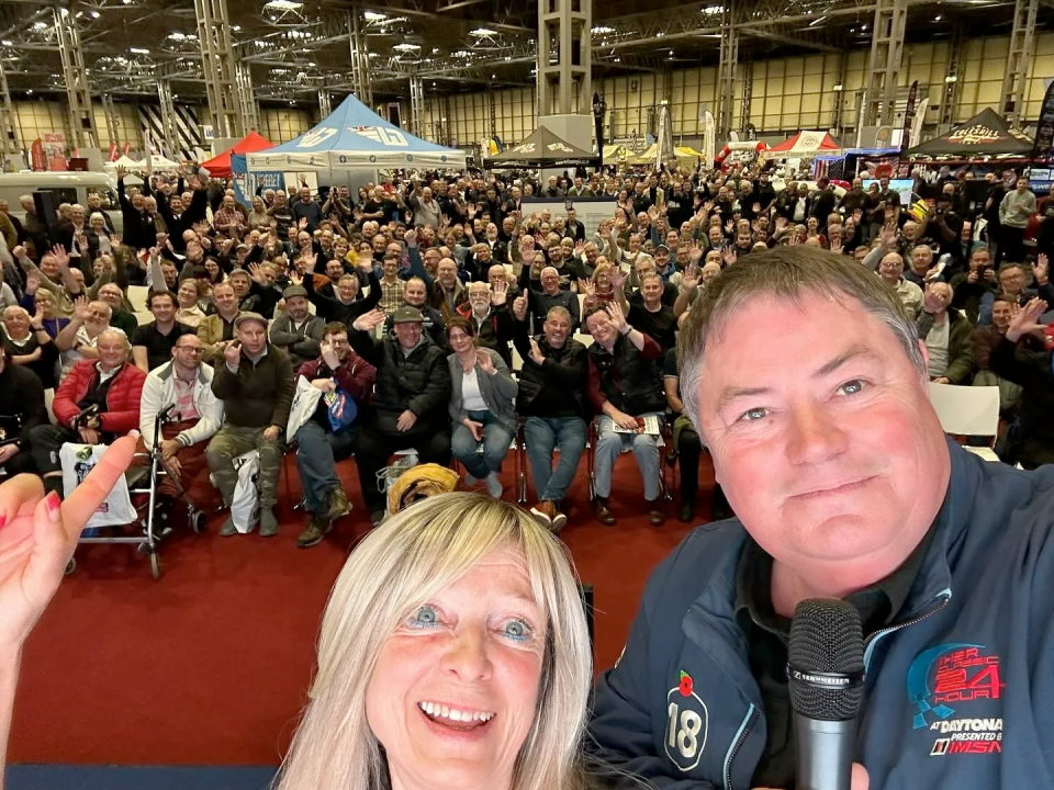 Two people in the foreground taking a selfie with a large crowd waving in the background at an indoor event.