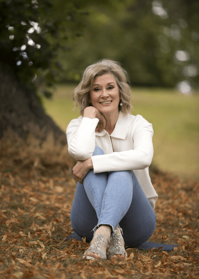 Woman sitting on the ground with fallen leaves, wearing jeans and a white cardigan.