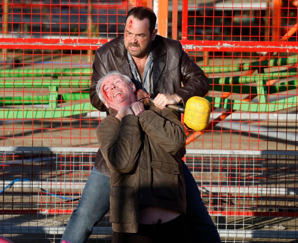 Danny Dyer in a brown leather jacket holds co-star Ned Dennehy in a headlock, a yellow mallet held in his left hand, as they film a fight scene.