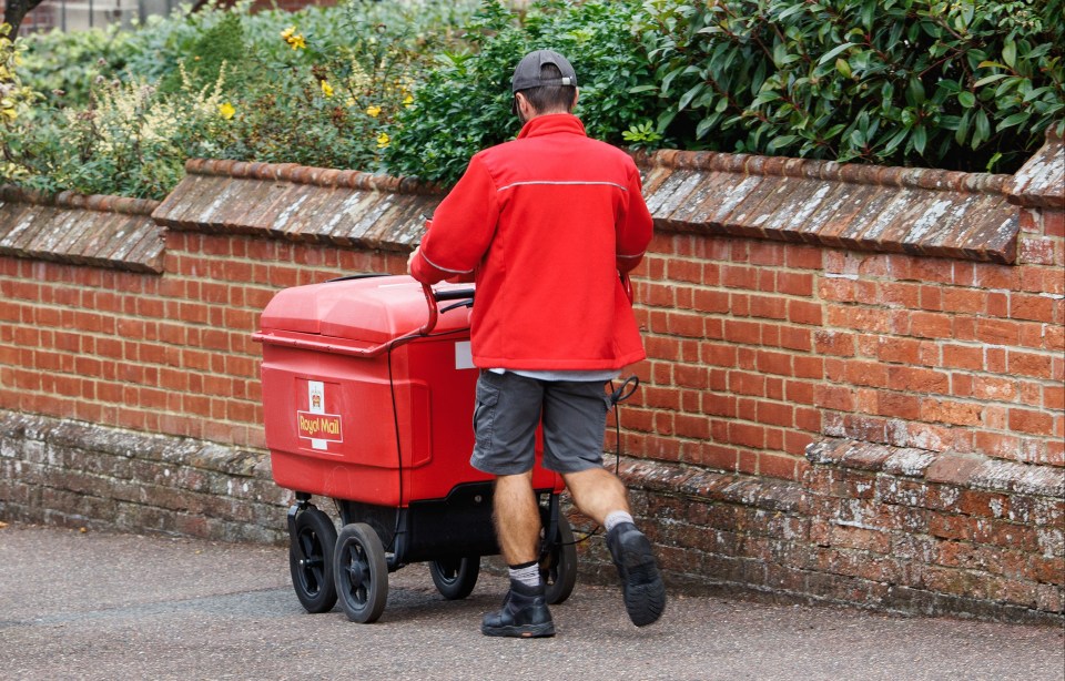 A Royal Mail delivery worker in a red jacket pushes a red mail cart along a sidewalk next to a brick wall.