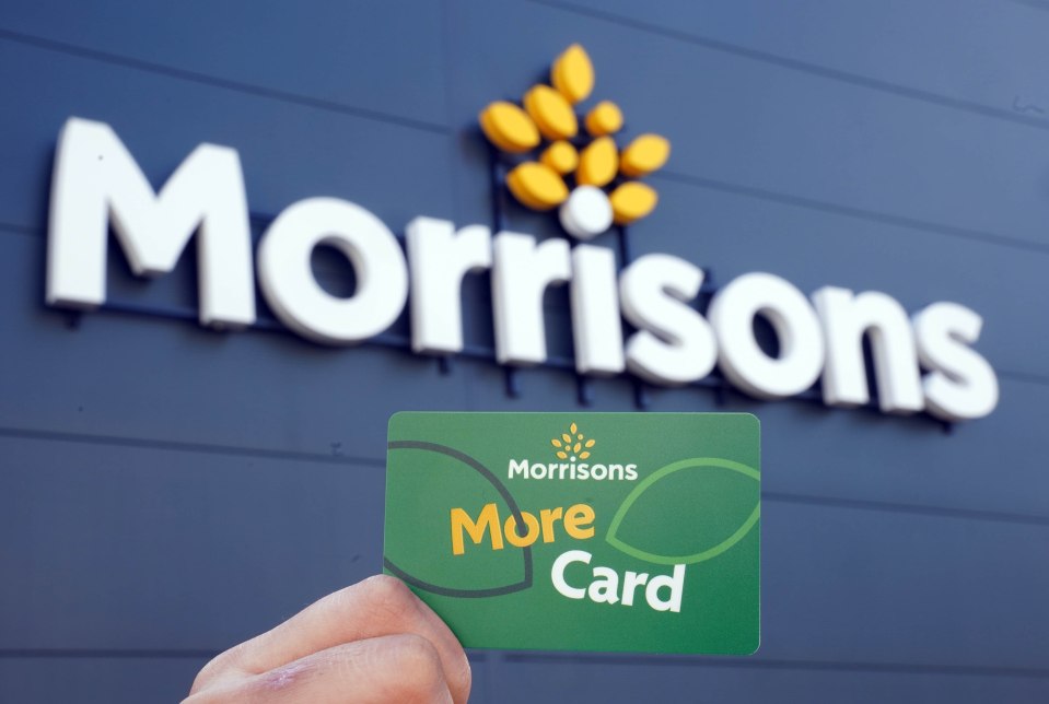 Hand holding a Morrisons More Card in front of a Morrisons store sign.
