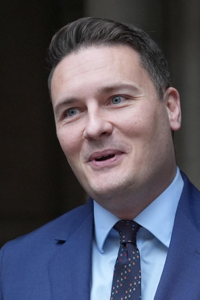Wes Streeting looking up and to the right, wearing a blue suit, light blue shirt, and dark blue patterned tie.