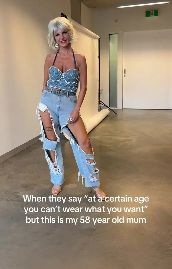 A 58-year-old woman with short blonde hair and earrings, wearing a pearl-studded denim crop top, distressed ripped jeans, and clear heels, standing in a studio with a white backdrop.