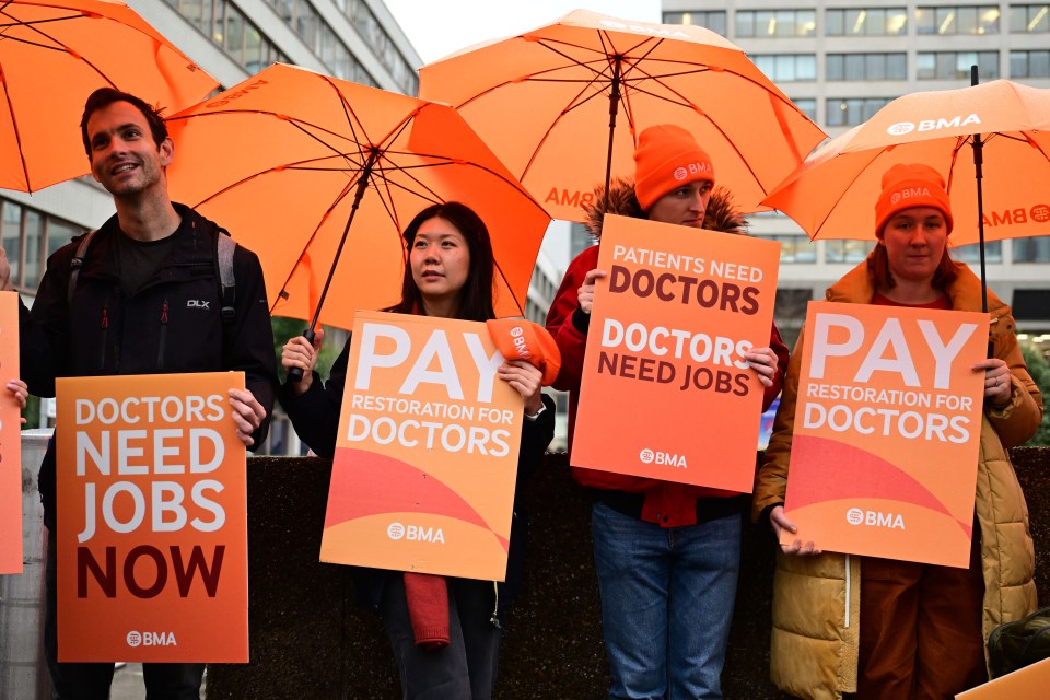 Four doctors hold protest signs about pay and jobs, and orange umbrellas.