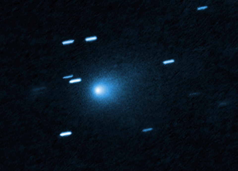 Hubble image of interstellar comet 3I/ATLAS, a bright blue-white point of light, surrounded by smaller, streaked objects, against a dark blue background.
