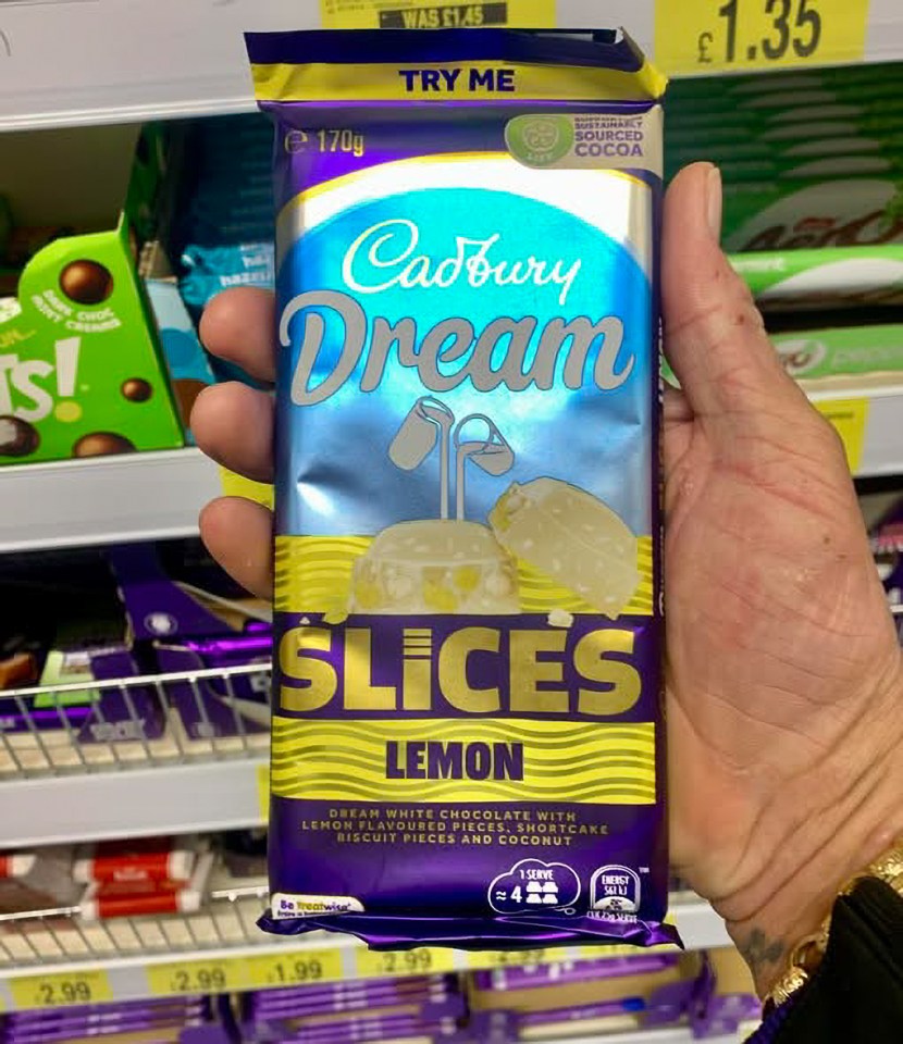 A hand holding a purple and yellow Cadbury Dream Slices Lemon chocolate bar in a store aisle.