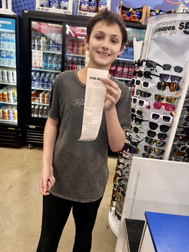 A smiling boy holding up a long receipt from "Five Below" in front of a store display of sunglasses and a refrigerator case of drinks.
