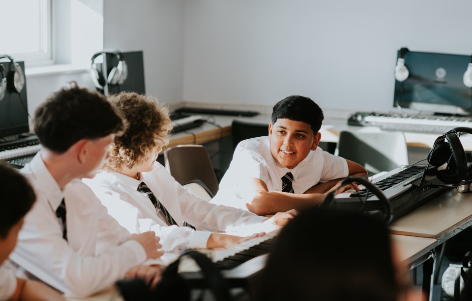 Four high school boys in uniform chat while sitting in a row in a sunny music classroom.