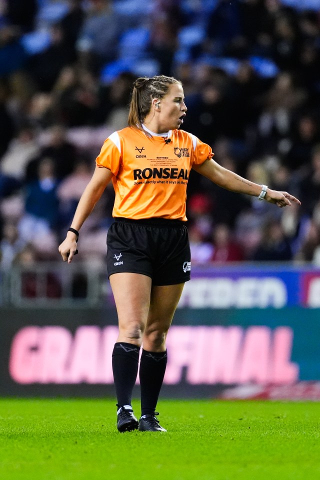 Tara, a St Helens player, officiating a Wigan Warriors versus St Helens rugby match in an orange referee shirt, black shorts, and black socks, with a crowd and "GRAND FINAL" sign in the blurred background.