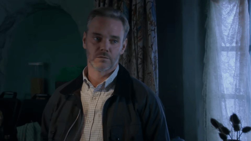 A man with short grey hair and a beard, wearing a dark jacket over a plaid shirt, looks to the right with a neutral expression.