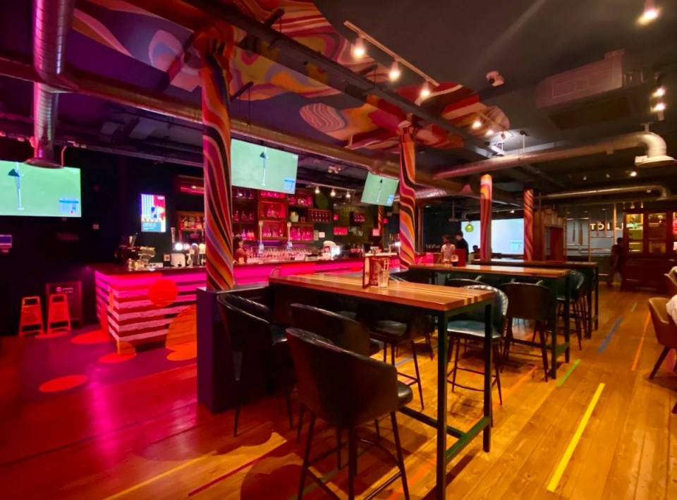 Interior of a modern bar with pink lighting, a colorful ceiling, and high-top tables.
