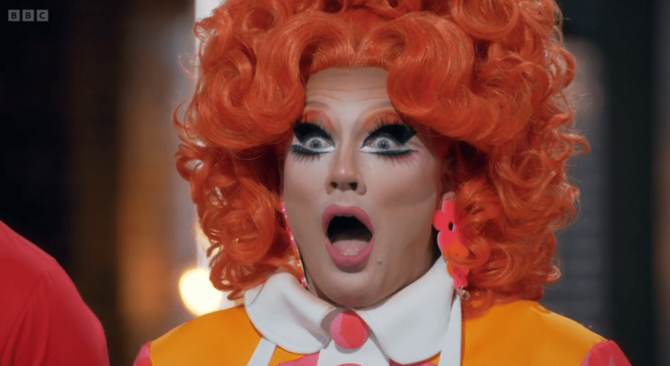 A drag queen with orange hair and makeup looks surprised with an open mouth.