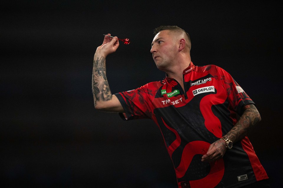 A dart player in a red and black shirt with tattoos on both arms throwing a dart.