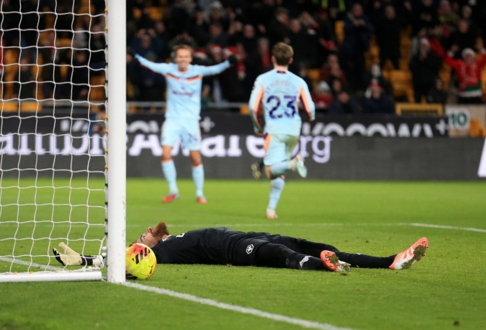 A goalkeeper lies on the ground next to a goalpost, while two players celebrate a goal behind him during a football match.