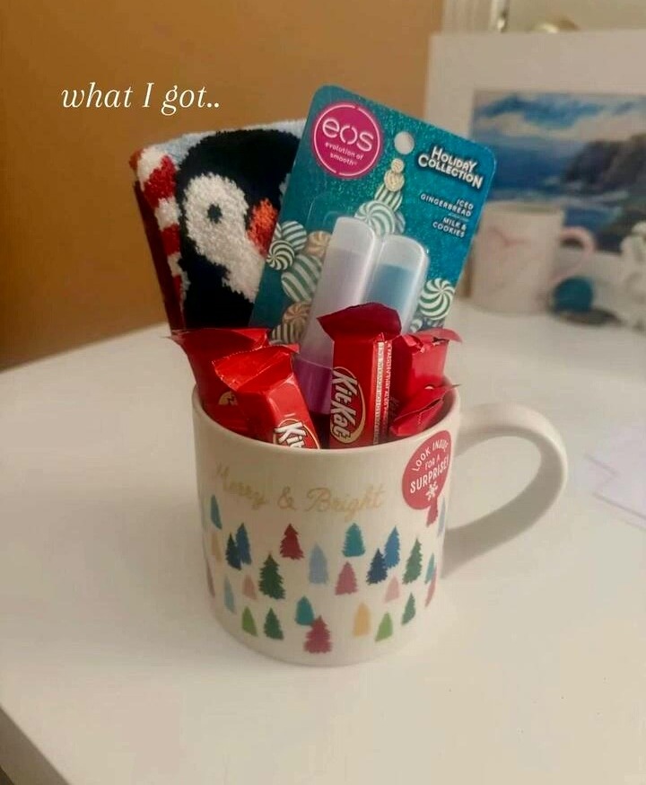 A white mug with "Merry & Bright" and colorful Christmas trees printed on it, filled with a pair of penguin socks, an EOS Holiday Collection lip balm set, and Kit Kat chocolate bars.