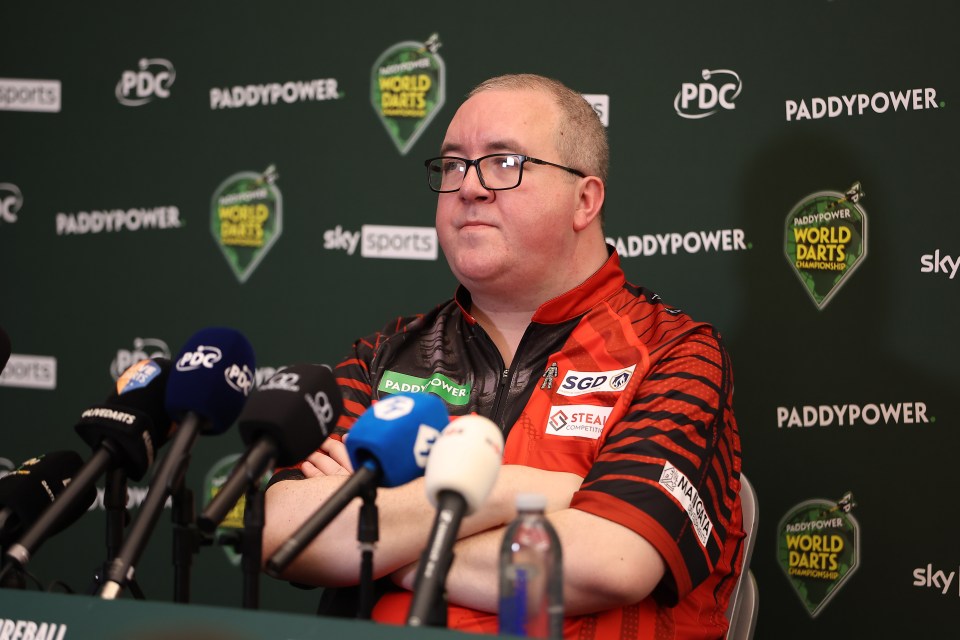 Stephen Bunting in a red and black shirt at a press conference with microphones in front of him and a "Paddy Power World Darts Championship" banner behind him.