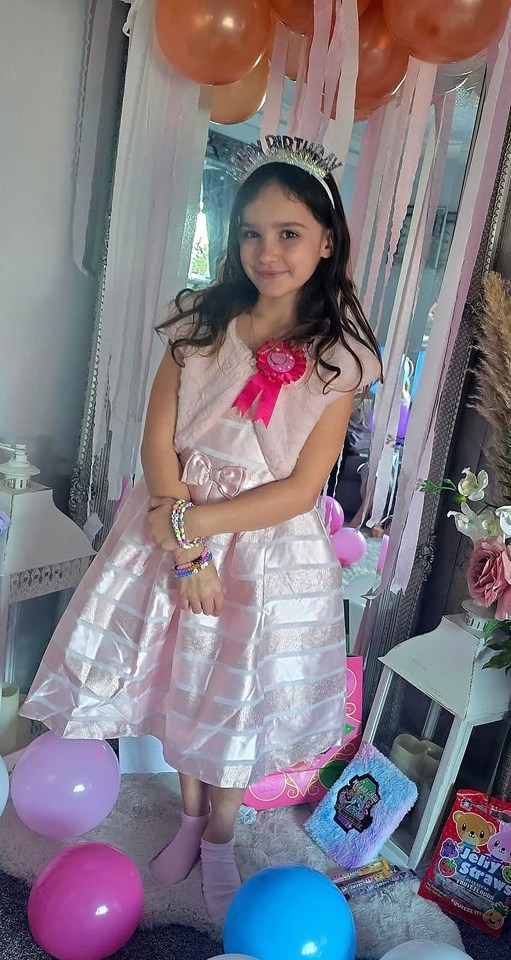 Aria Thorpe, a young girl, smiles while wearing a pink dress, a birthday crown, and colorful bracelets.
