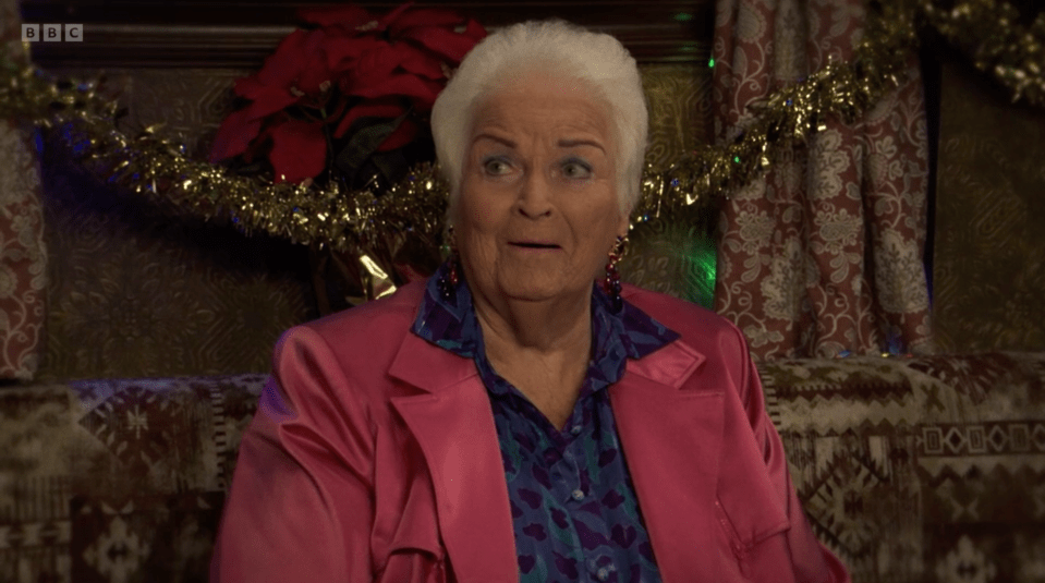 An elderly woman with white hair wearing a pink blazer, blue patterned shirt, and red earrings is seated on a patterned sofa.
