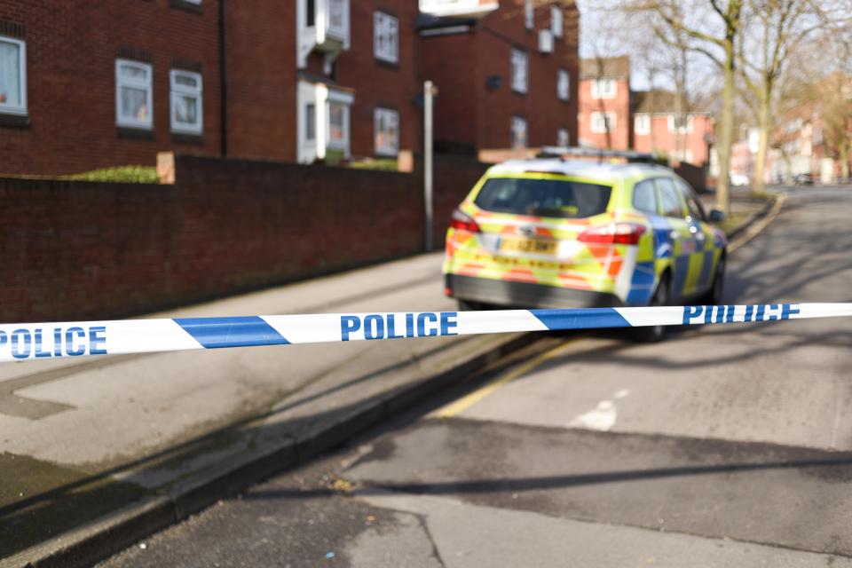Police tape blocking off a road with a police car and buildings in the background.