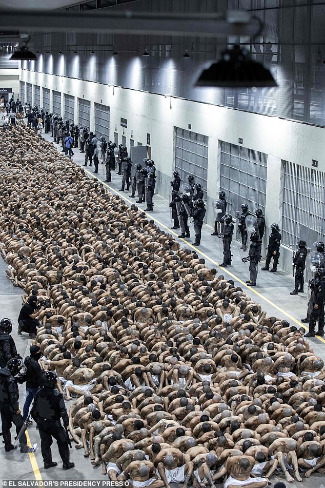Hundreds of rows of prisoners are watched over with their heads down by visor-wearing armed guards