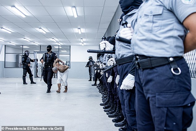 Guards armed with batons watch on as a prisoner is led through the prison by an officer carrying a large gun