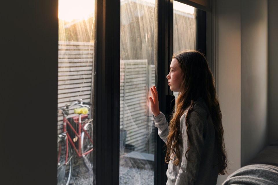Teenage girl looking through a glass sliding door with her hand resting on the glass, appearing contemplative.