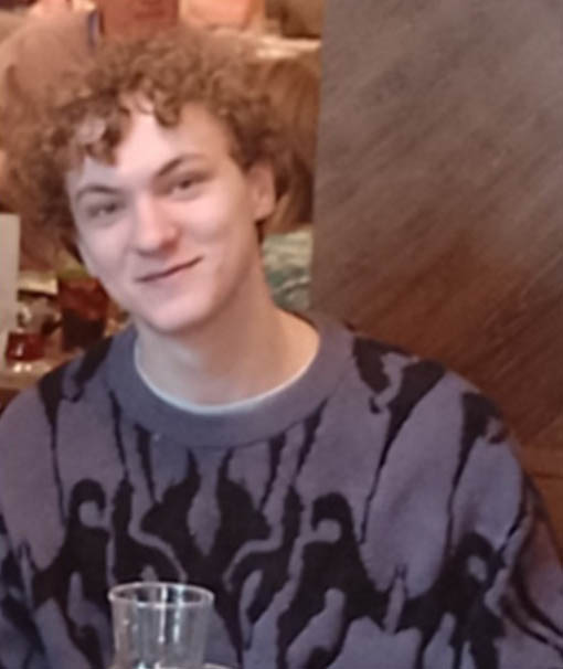 Adam, a 19-year-old with curly brown hair, wearing a purple and black patterned sweater.