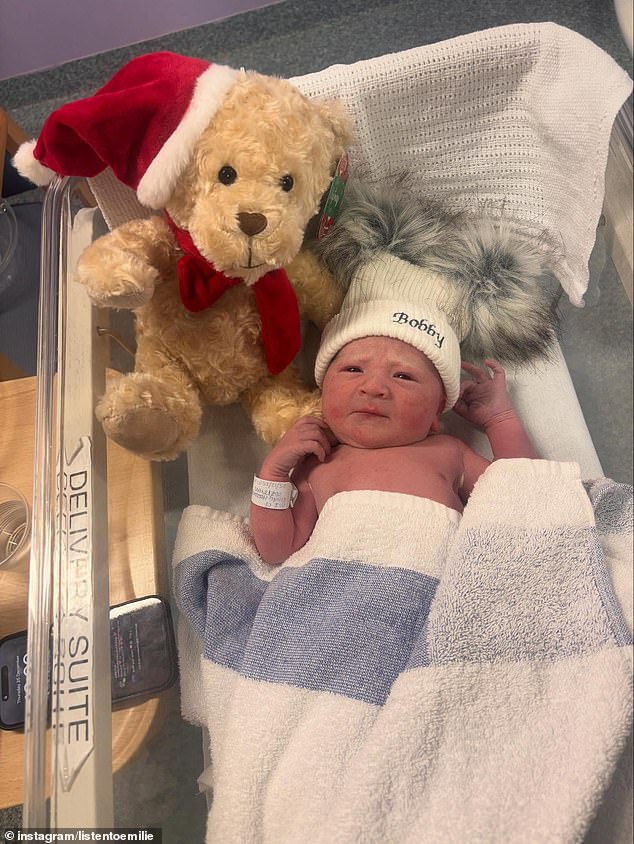 The beautiful newborn can be seen with a personalised beanie as he lays in his hospital cot next to a teddy bear with a Christmas hat