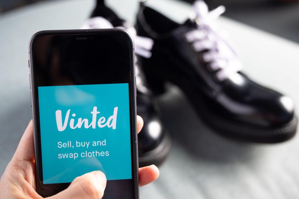 A hand holding an iPhone displaying the Vinted logo and slogan "Sell, buy and swap clothes," with a pair of black dress shoes in the background.