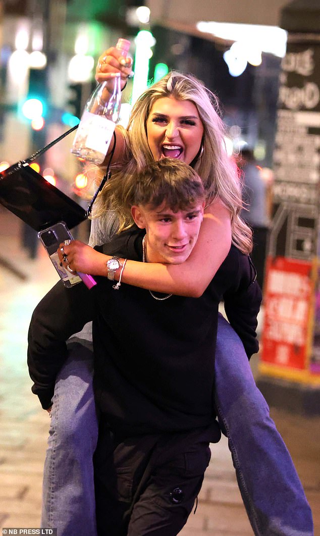 One young man gives a lady a piggy back as she raises a bottle of wine into the air in Leeds