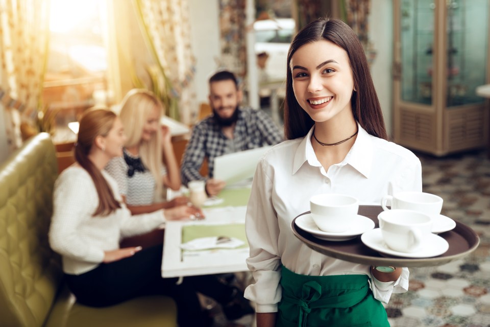 A smiling young waitress holding a tray of coffee cups in a cafe with customers seated at tables in the background.