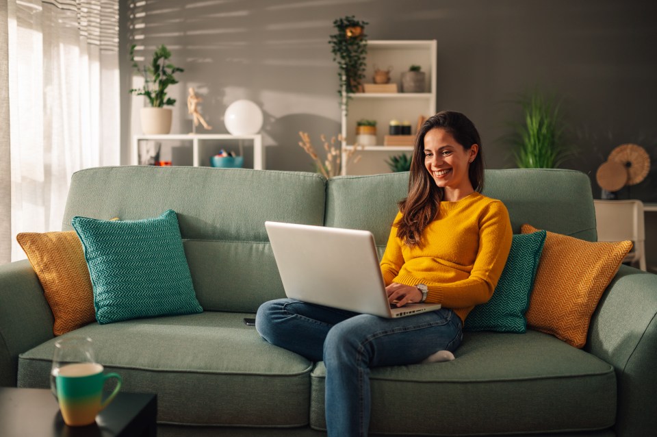 A woman smiling while working on her laptop from a mint couch.