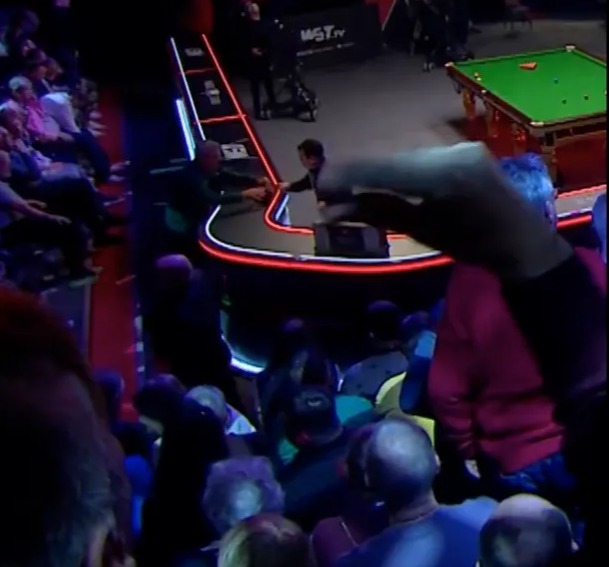 A snooker match in progress with players at a lit table, surrounded by spectators.