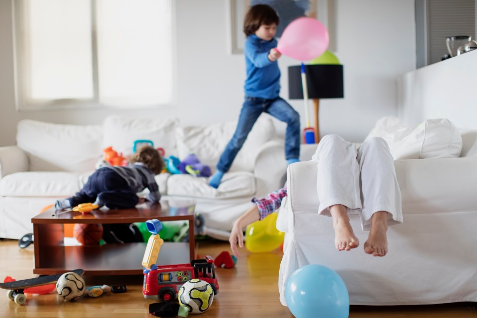 Two boys play in a messy living room, jumping from sofa to sofa while their mother rests, with only her feet visible.