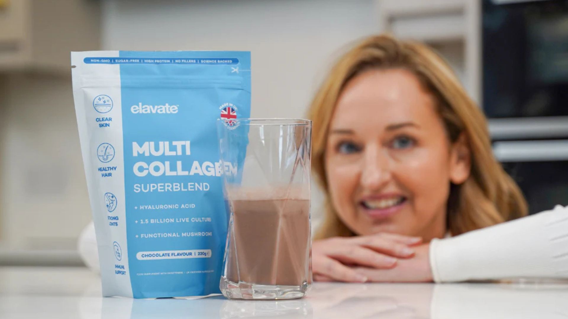 A smiling woman in the background, out of focus, looks at a bag of Elavate Multi Collagen Superblend and a glass of the chocolate-flavored drink.