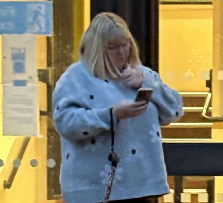 A blonde woman in a light blue sweater with dark spots looks at her phone.