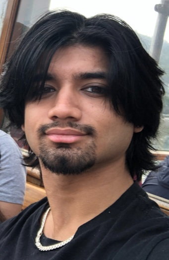 Aryan Sharma, a 20-year-old student, with dark hair and a beard, wearing a black shirt and silver chain.