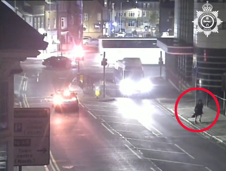 CCTV footage of a street at night, with a person in a black coat circled in red on the right sidewalk.