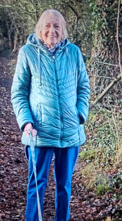 An older woman in a blue jacket holding a dog leash while standing on a leaf-covered path.