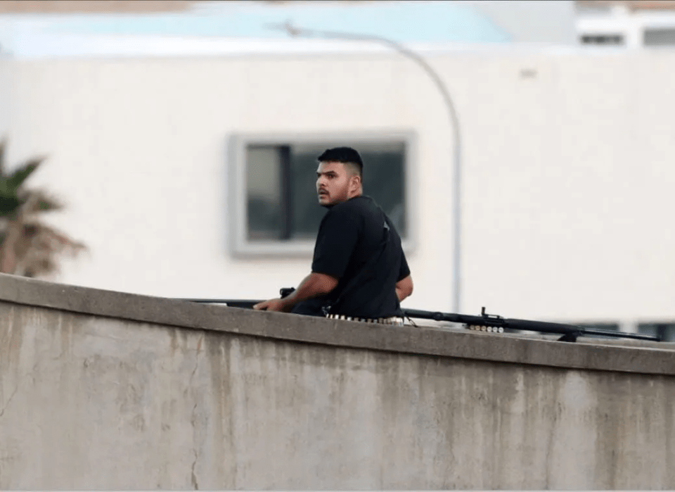 A man in a black shirt sitting behind a concrete wall with a long firearm, looking over his shoulder.