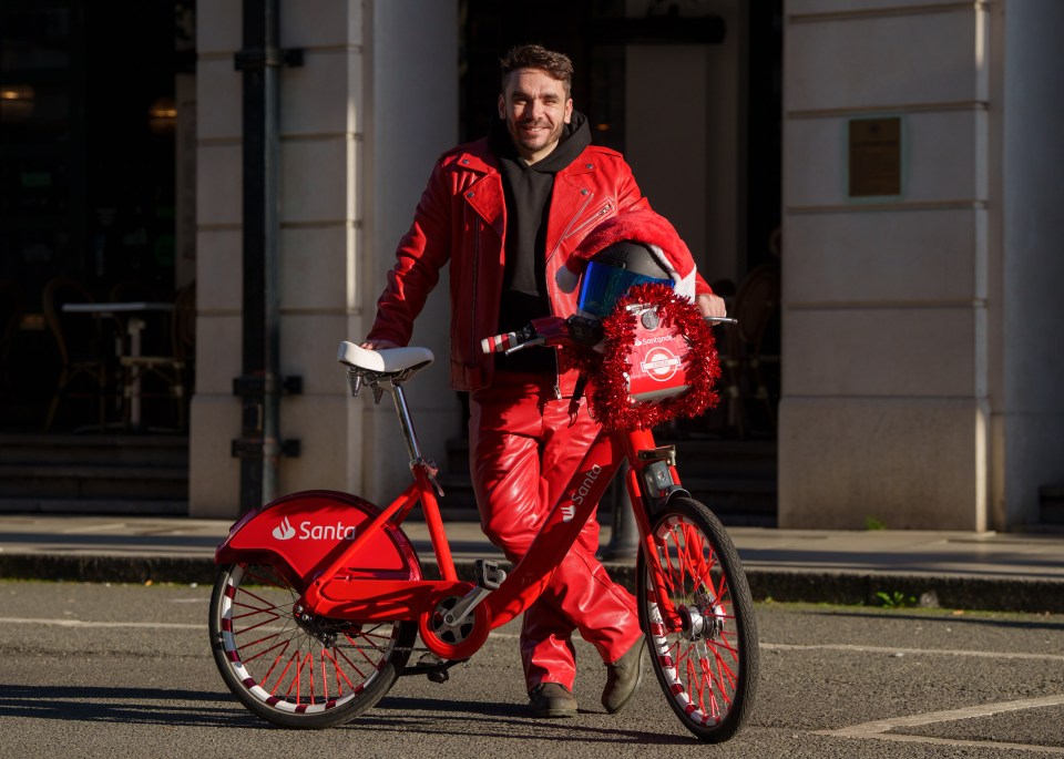 HarveyRidesBikes, in a red leather suit, stands next to a Santa-themed Santander Cycle with a decorated basket.